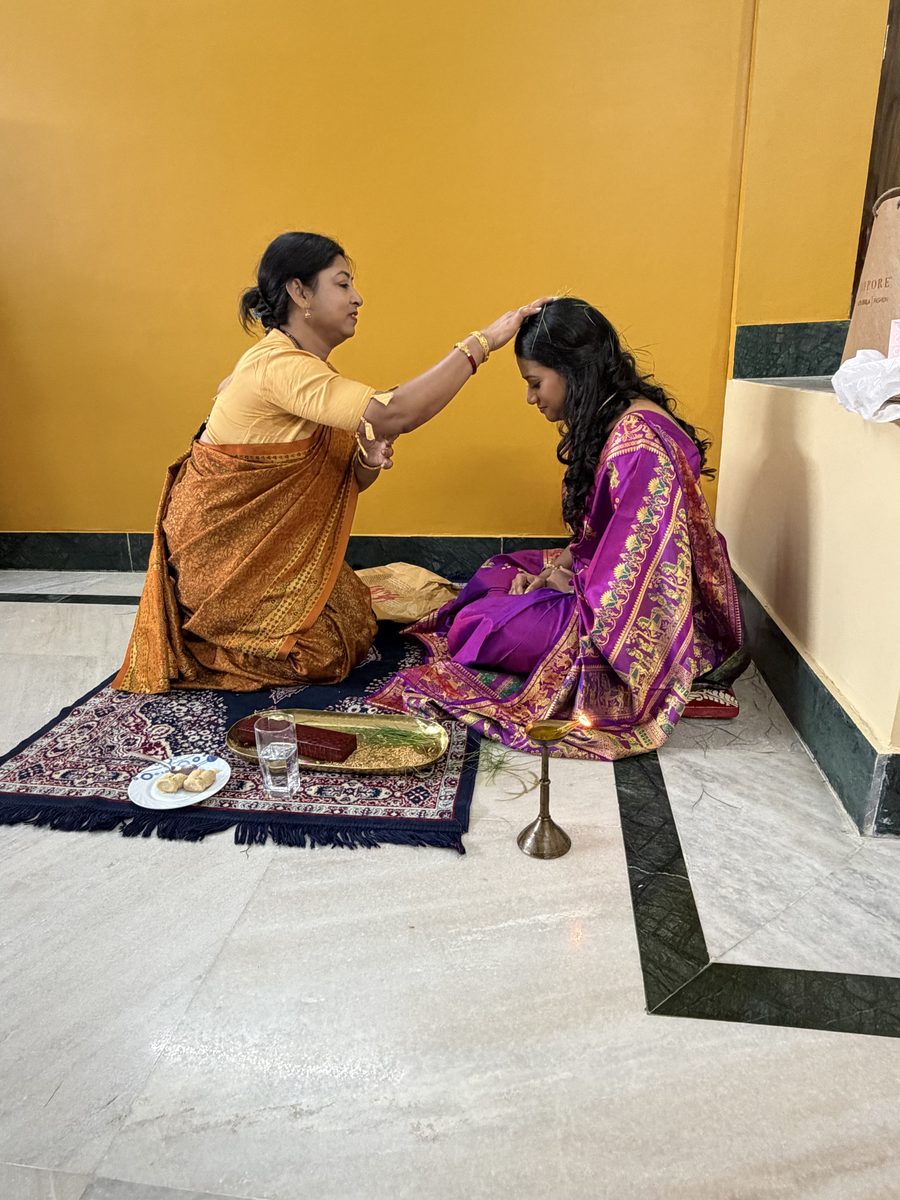 Woman in gold sari blessing Rituparna seated on the prayer mat