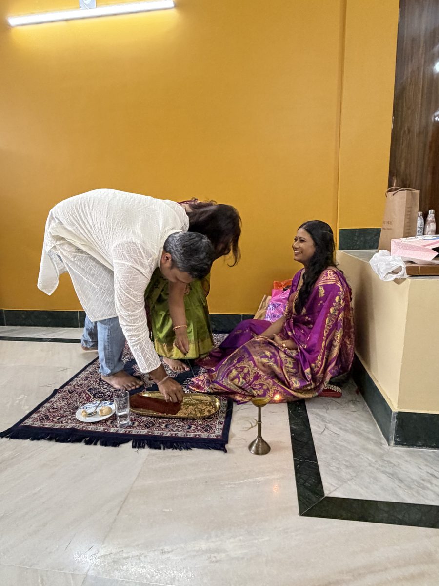 Woman in white clothing bending to bless Rituparna on the prayer mat